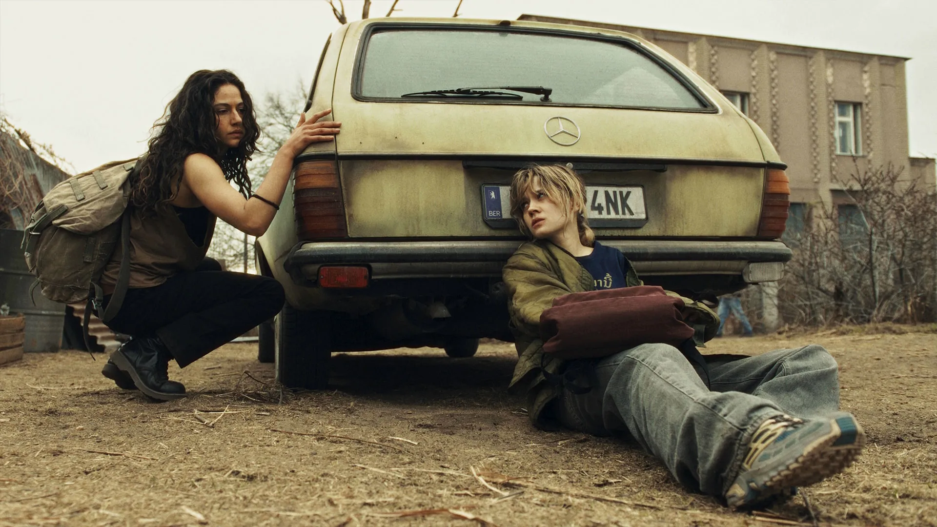 Two people shelter behind an older model car in a dusty outdoor area, with backpacks and buildings visible in the background.