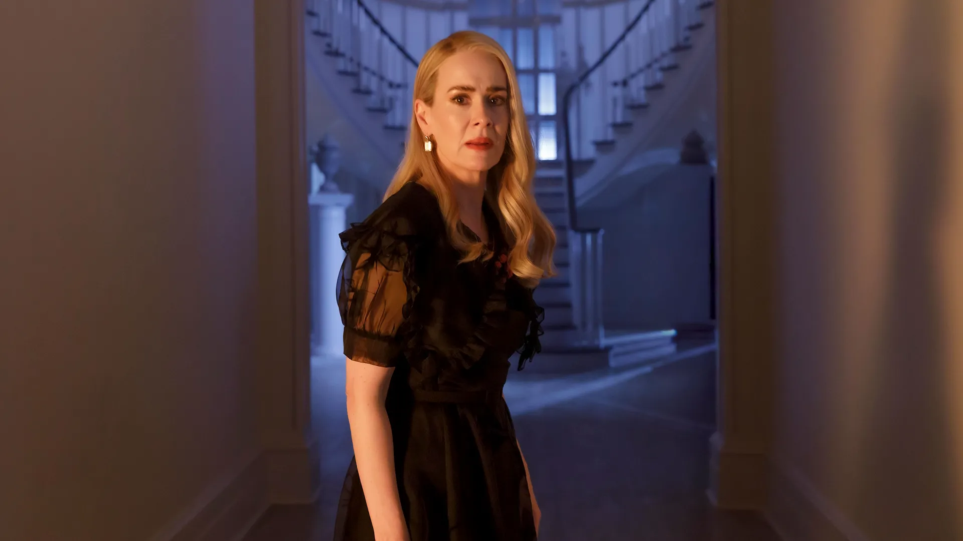 A blonde woman with a concerned expression looks back over her shoulder in a brightly lit hallway, wearing a black dress with sheer ruffled sleeves against the backdrop of a grand staircase.
