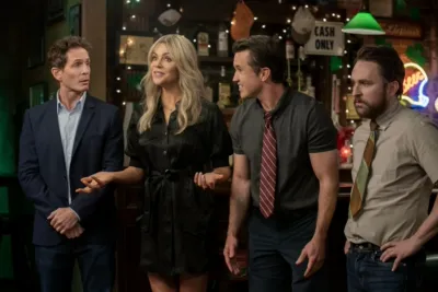 In a dimly lit college lecture hall with orange stadium seating, Dennis Reynolds (Glenn Howerton) wearing a brown corduroy button-down shirt and a canvas messenger bag stands next to Dee Reynolds (Kaitlin Olson) wearing a graphic t-shirt and a blue varsity jacket as they look ahead with expressions of mild concern and confusion.