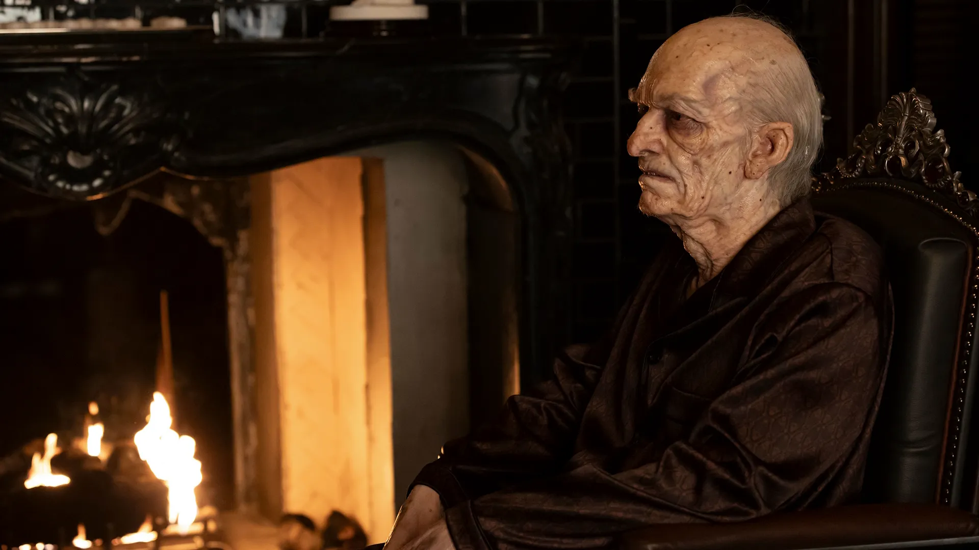 An elderly, frail man with heavily wrinkled, pale skin sits in a dark leather armchair next to a roaring fireplace, wearing a dark maroon silk robe or pajamas.