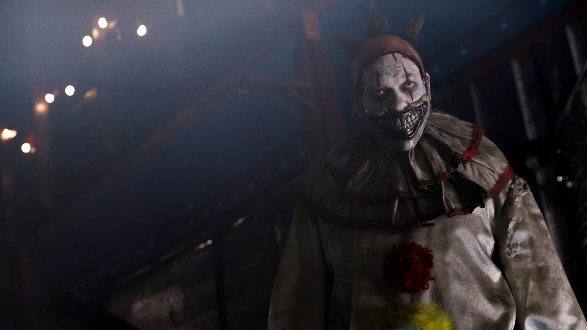 A terrifying clown with a distorted, oversized grin and pale, dirty makeup stares menacingly forward while standing under dim, foggy stage lights.