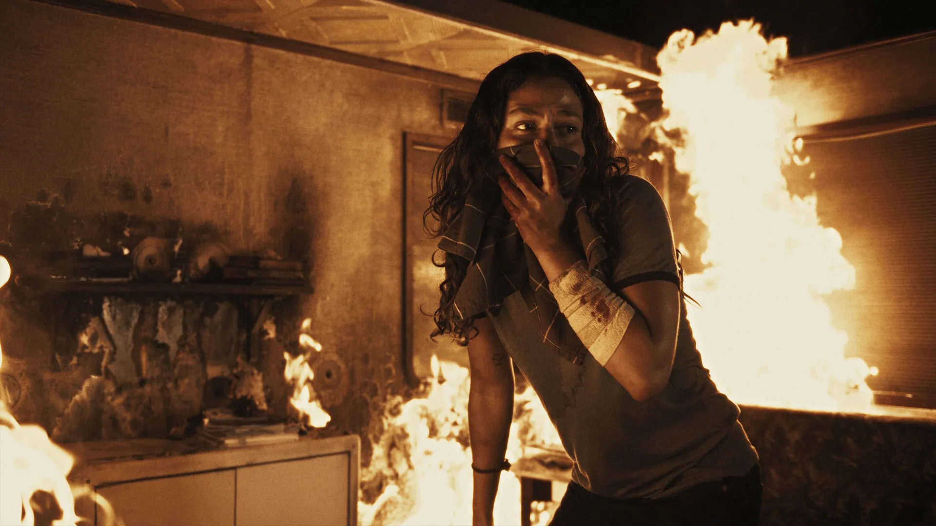 Soma Pysall as Phoebe covers their mouth with a piece of fabric while surrounded by flames inside a burning room.