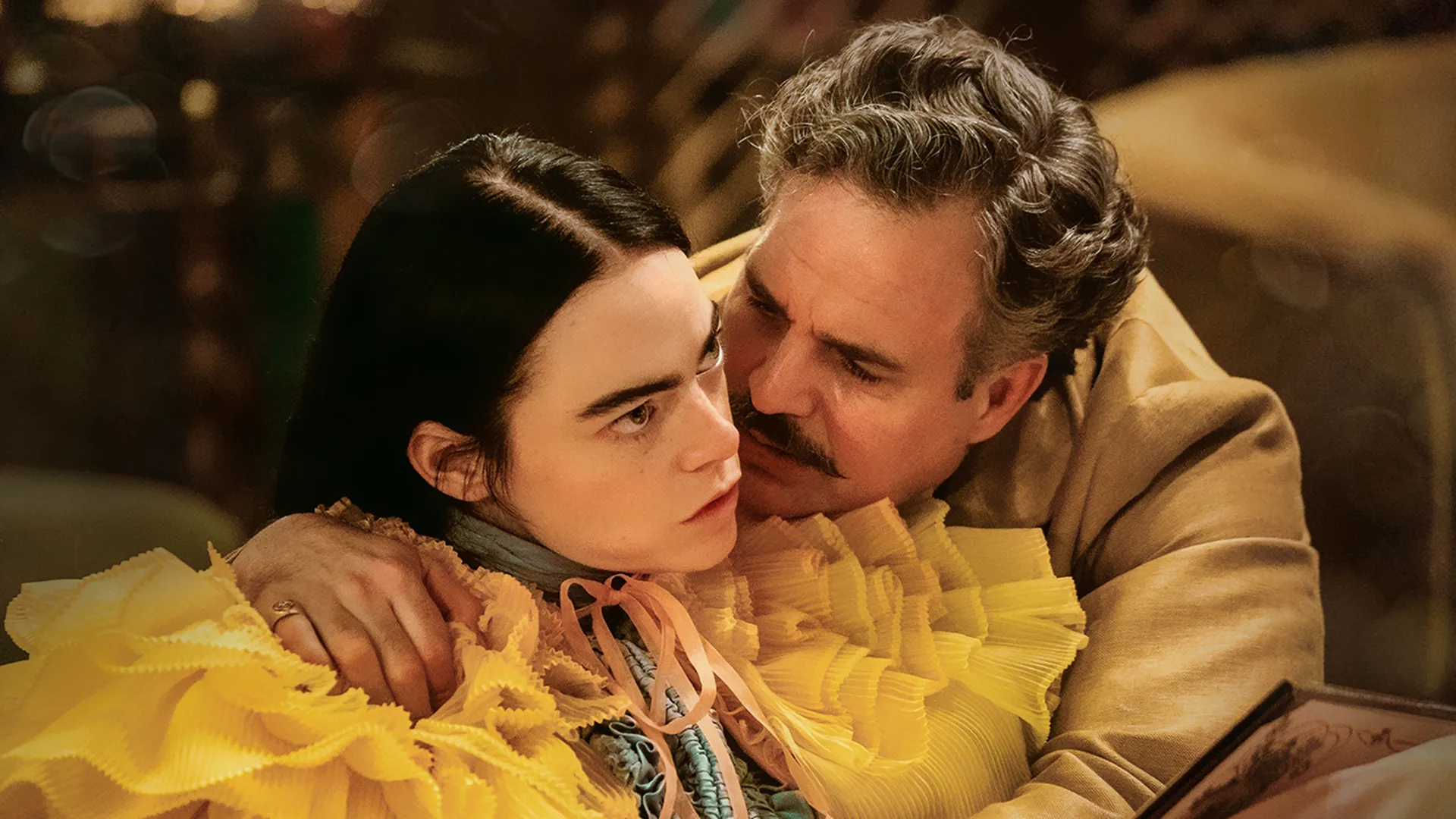 Emma Stone as Bella Baxter stares blankly forward in an ornate yellow ruffled dress while Mark Ruffalo as Duncan Wedderburn embraces her from behind, whispering in her ear in a scene from Poor Things.