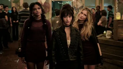 Sasha (Jordan Leftwich), Briar (Malina Pauli Weissman), and Tegan (Ora Duplass) stand side-by-side in dark, witchy attire amidst a crowded room decorated with string lights in a scene from the supernatural drama series Coven Academy.