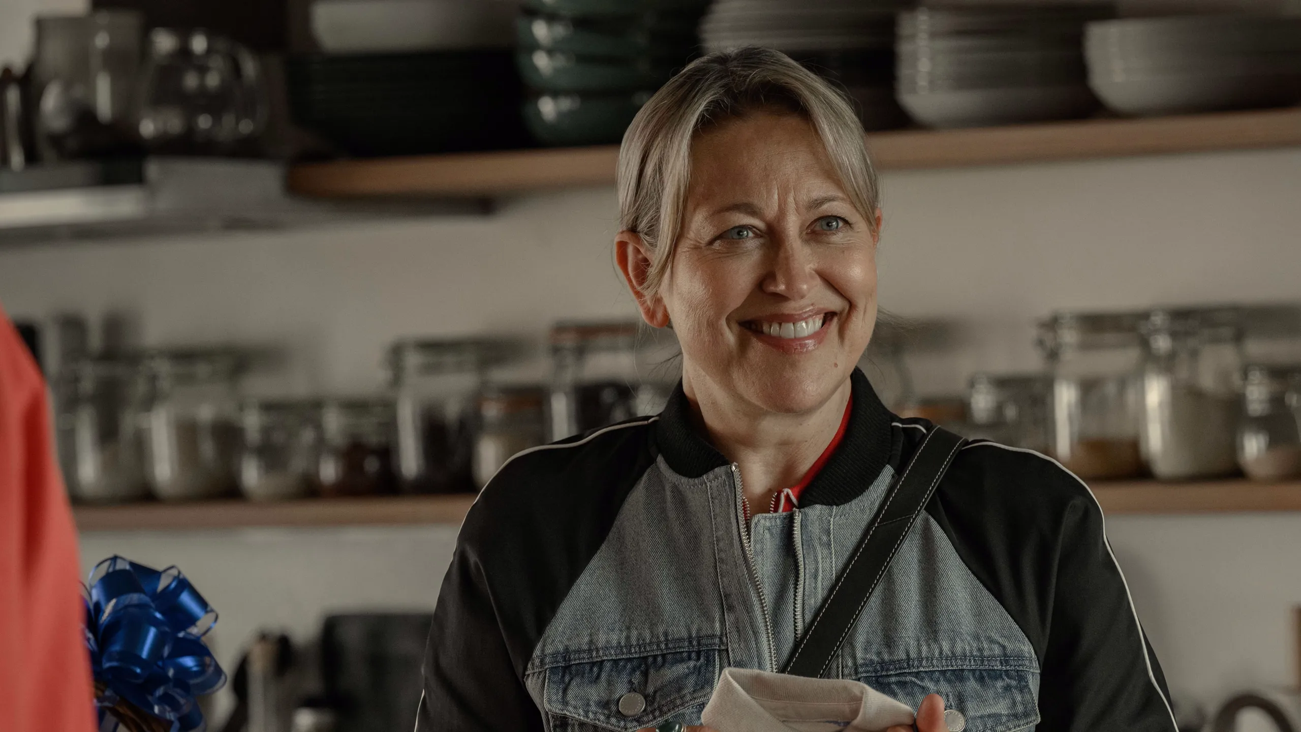 Alice stands in a kitchen holding a dish towel, with shelves of kitchenware in the background.