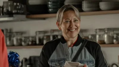 Alice stands in a kitchen holding a dish towel, with shelves of kitchenware in the background.