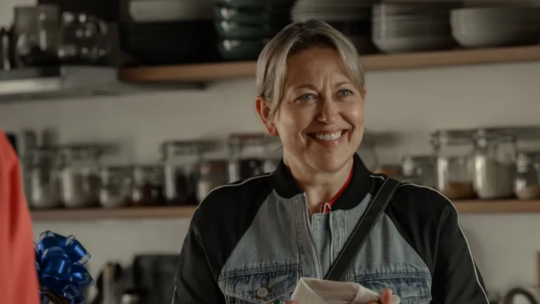 Alice stands in a kitchen holding a dish towel, with shelves of kitchenware in the background.