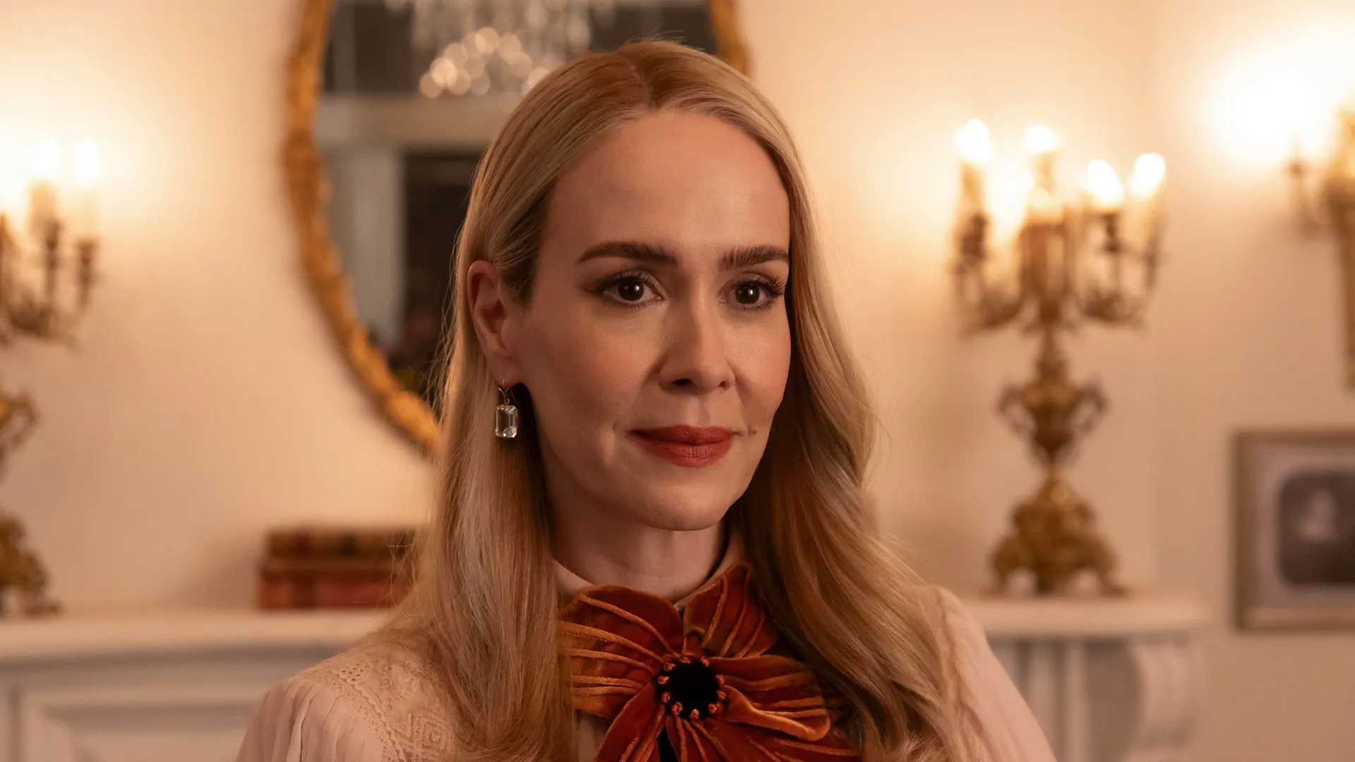 Sarah Paulson with styled blonde hair, wearing a soft pink blouse adorned with a large, elaborate orange velvet bow around her neck as she smiles gently in a warmly lit room.