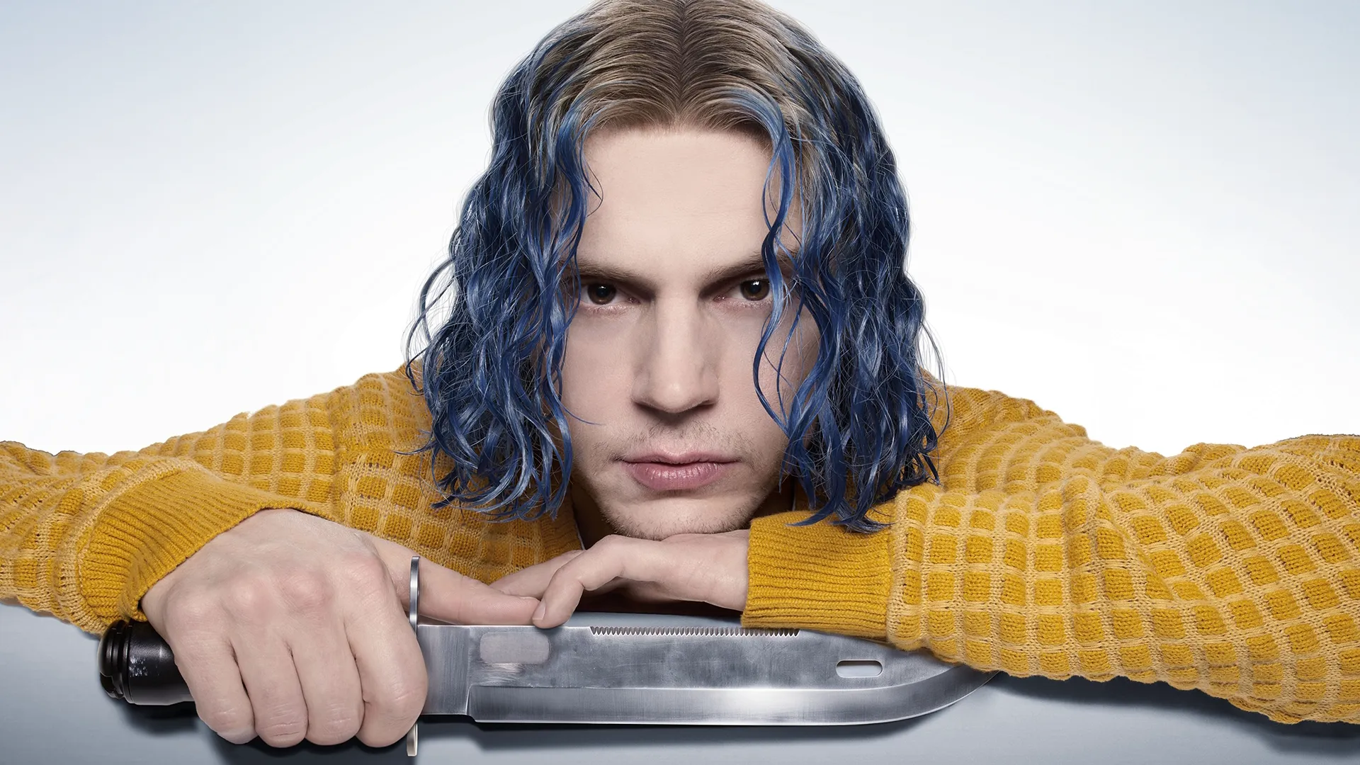 A man with wavy blue-streaked hair leans forward in a yellow textured sweater, resting his hands and chin over a large, metallic knife against a white background.