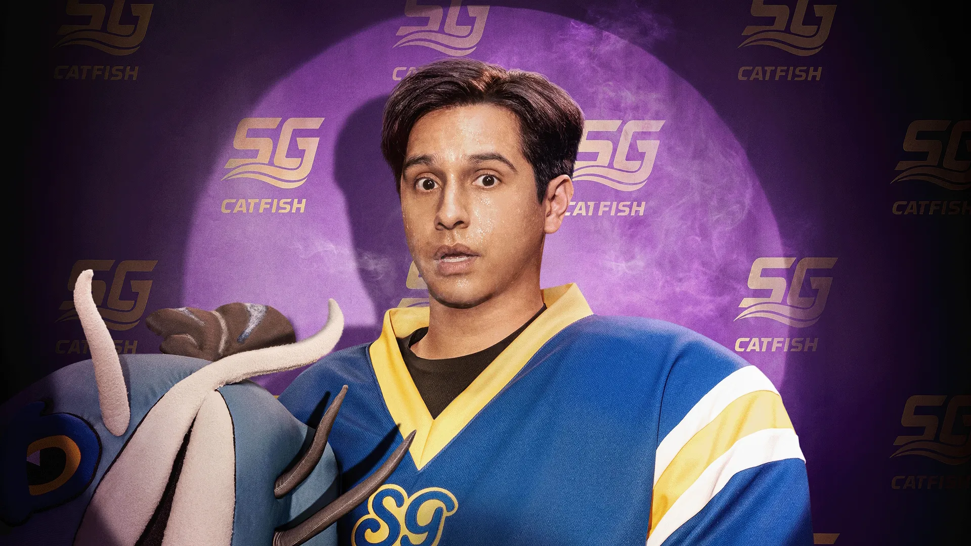 Danny Cruz (Frankie A. Rodriguez) parado, com cara de surpresa e suado, segurando uma cabeça de mascote azul em frente a um fundo roxo com o logotipo do time "SG CATFISH" estampado.