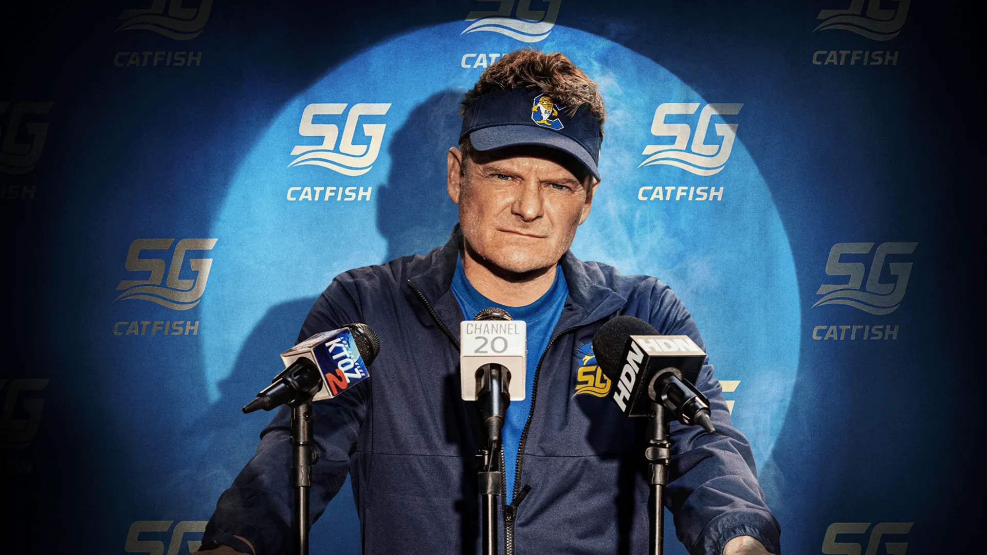 O técnico Jake Hudson (Steve Zahn) fala com uma expressão séria e intensa atrás de um pódio com microfones, vestindo uma jaqueta azul da equipe e uma viseira azul combinando com a inscrição "SG CATFISH", em frente a um fundo azul iluminado por holofotes.