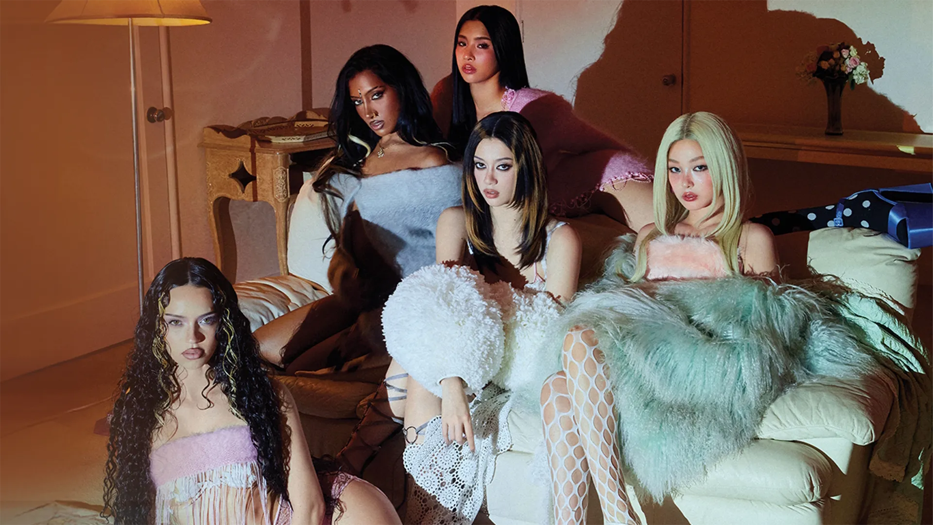 Five members of the girl group KATSEYE—Daniela, Lara, Yoonchae, Megan, and Sophia—pose close together on and around a white sofa in a warmly lit room with dramatic shadows casting against the wall behind them.