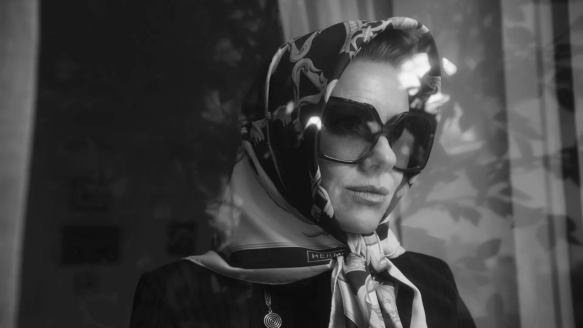 Jackie Kennedy Onassis (Naomi Watts) wearing large glasses and head covering in a black and white set photo from FX's Love Story.