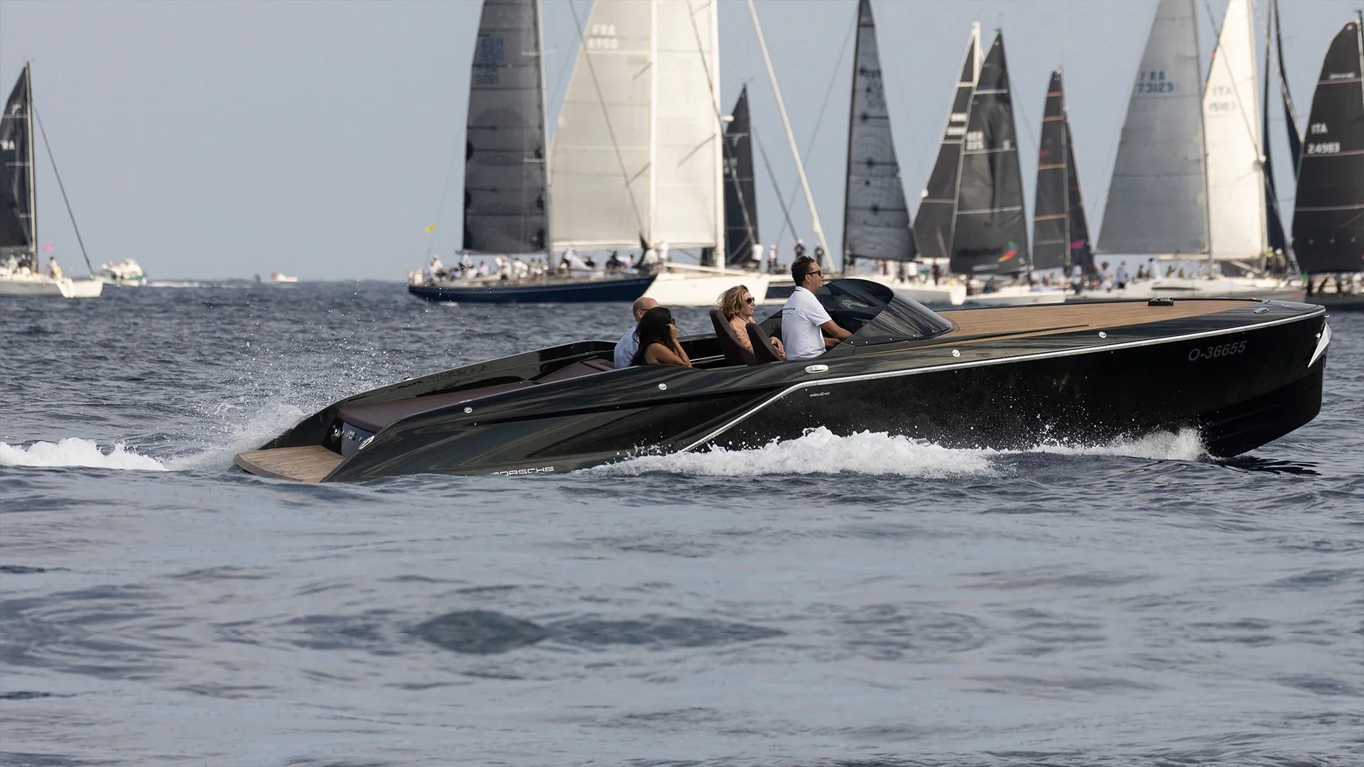 A luxury motorboat travels across open water with multiple sailing yachts in the background.