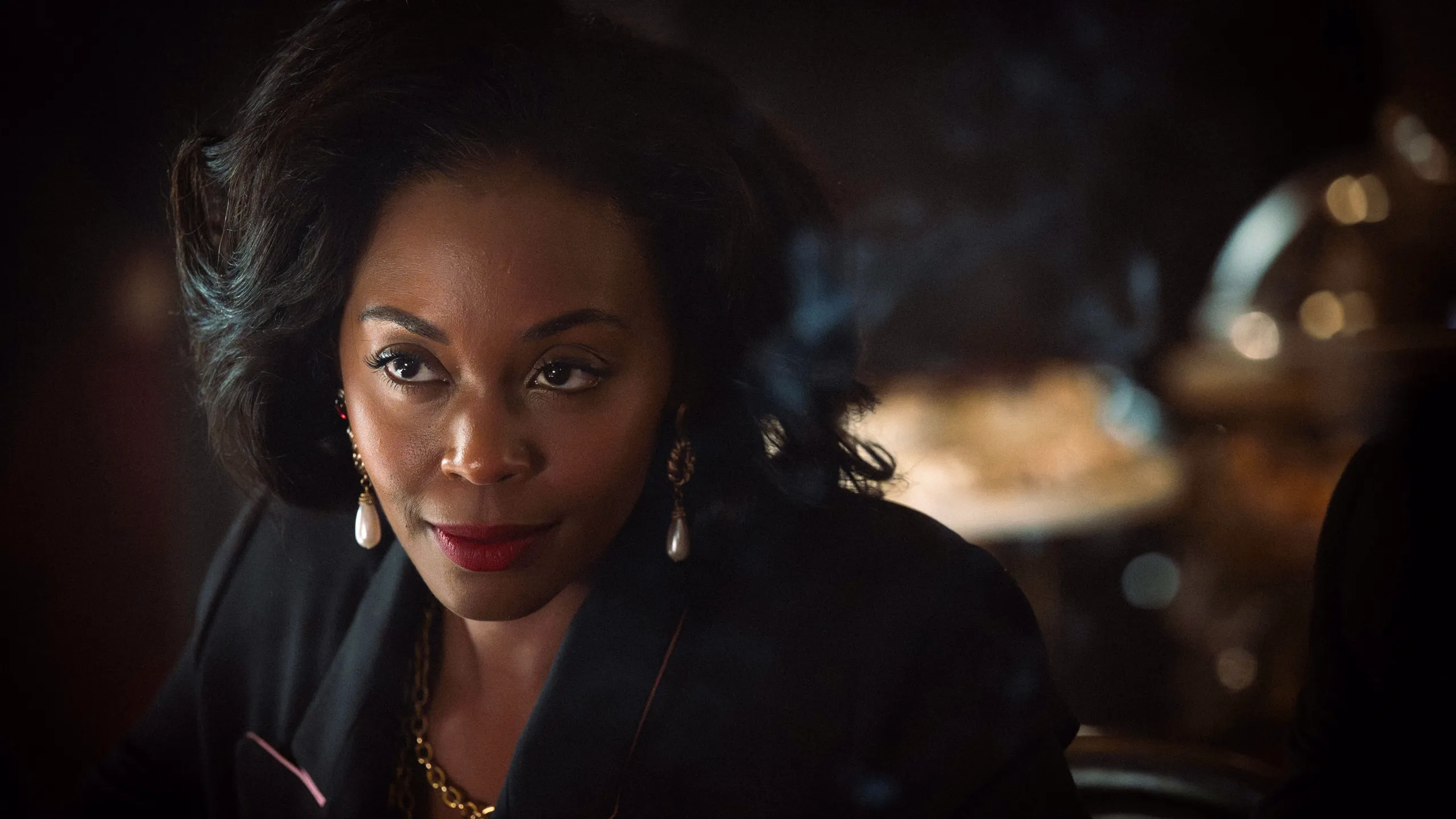 Cameron Cook (Nafessa Williams) in close-up wearing gold jewellery, lit in a dim interior setting in Rivals season 2.