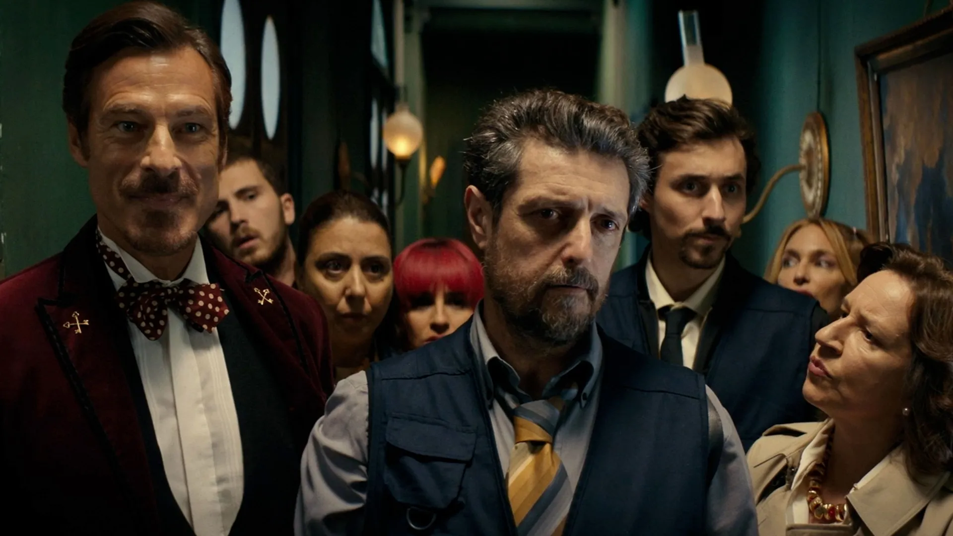 An ensemble cast of travelers, including actors Paulo Pires and Adriano Carvalho, looking suspiciously toward the camera in a narrow, green-walled hallway from the mystery series "Si Es Martes Es Asesinato."