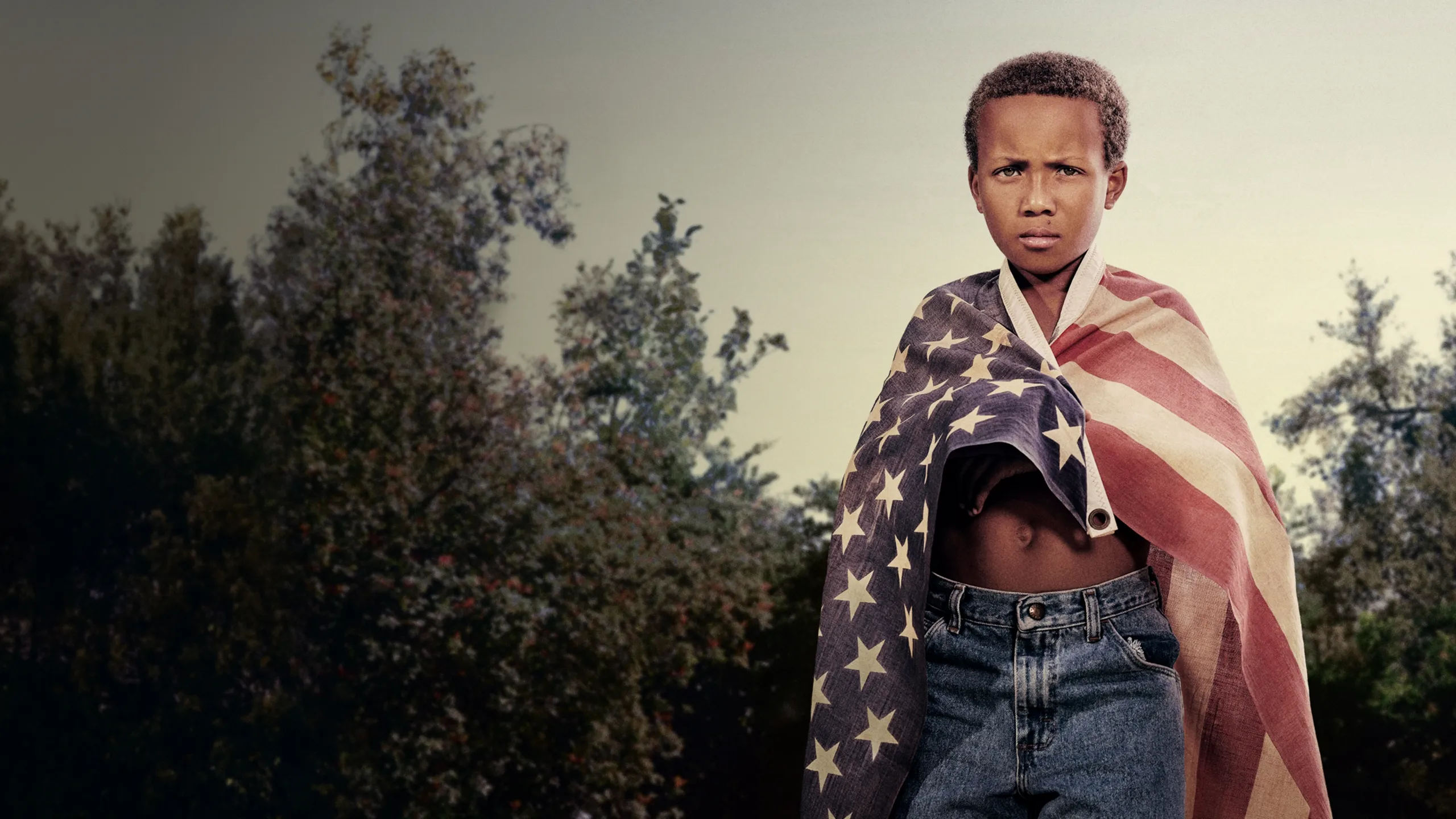 A man with locs holding a child in the air, an American flag in the background