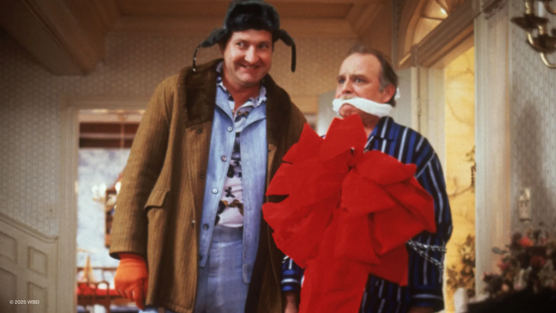 Cousin Eddie (Randy Quaid) smiles next to a terrified Frank Shirley (Brian Doyle-Murray), who is gagged and tied up with a large gift bow.