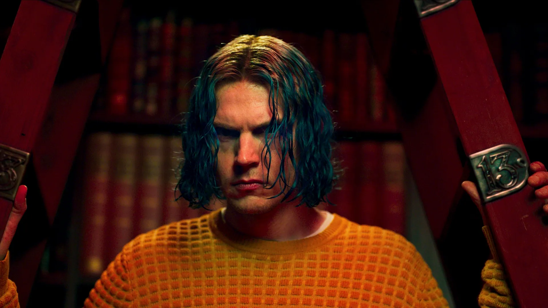 Kai Anderson (Evan Peters) glares forward with a tense, intense expression, framed by a red structure with a metal plate displaying the number 13, wearing an orange waffle-knit sweater and sporting shoulder-length blue hair with reddish roots against a backdrop of bookshelves.