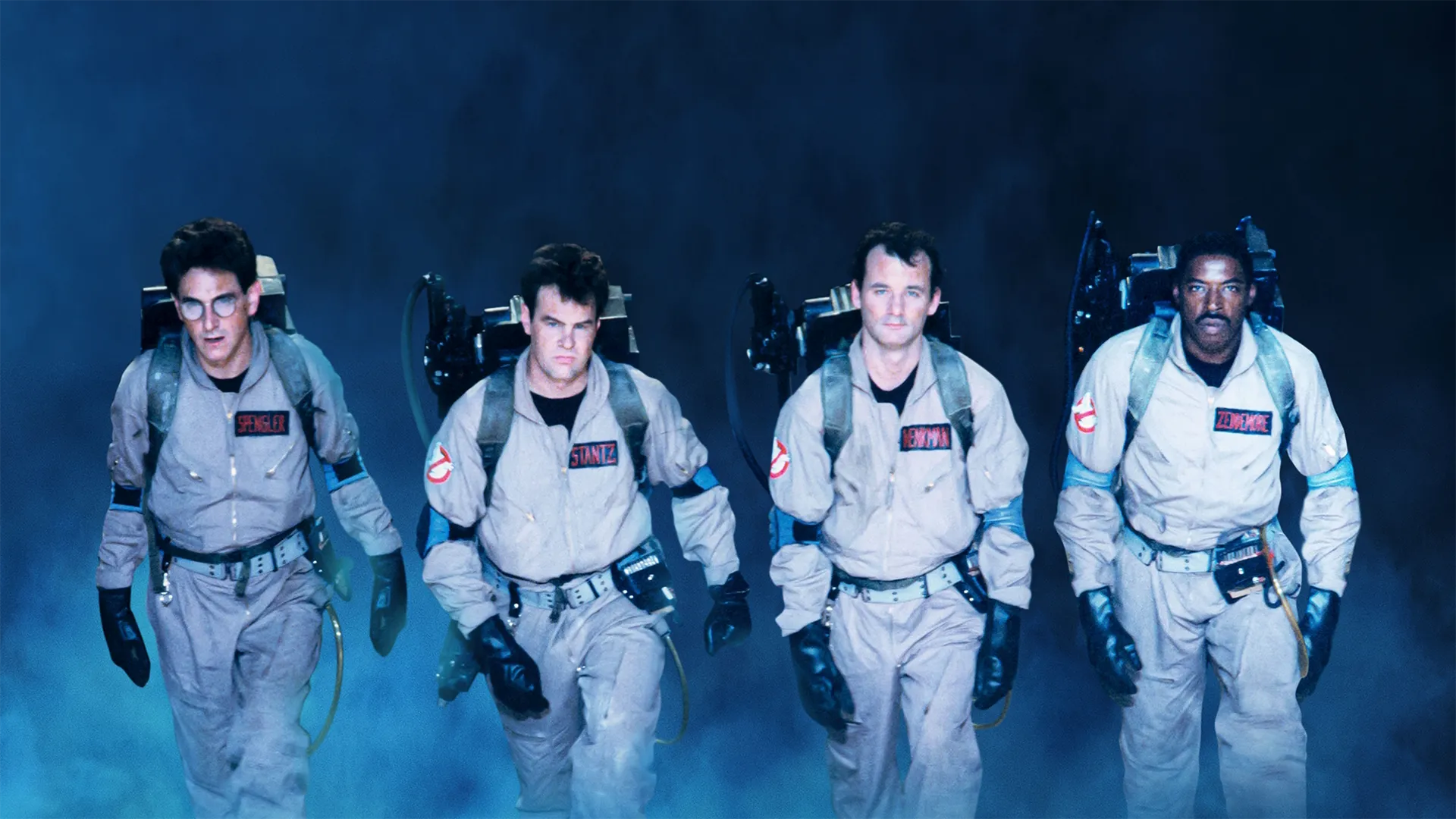 An iconic promotional image from Ghostbusters (1984) shows the four main characters, Egon Spengler, Ray Stantz, Peter Venkman, and Winston Zeddemore, walking forward side-by-side in their flight suits and proton packs through a misty, blue-lit atmosphere.