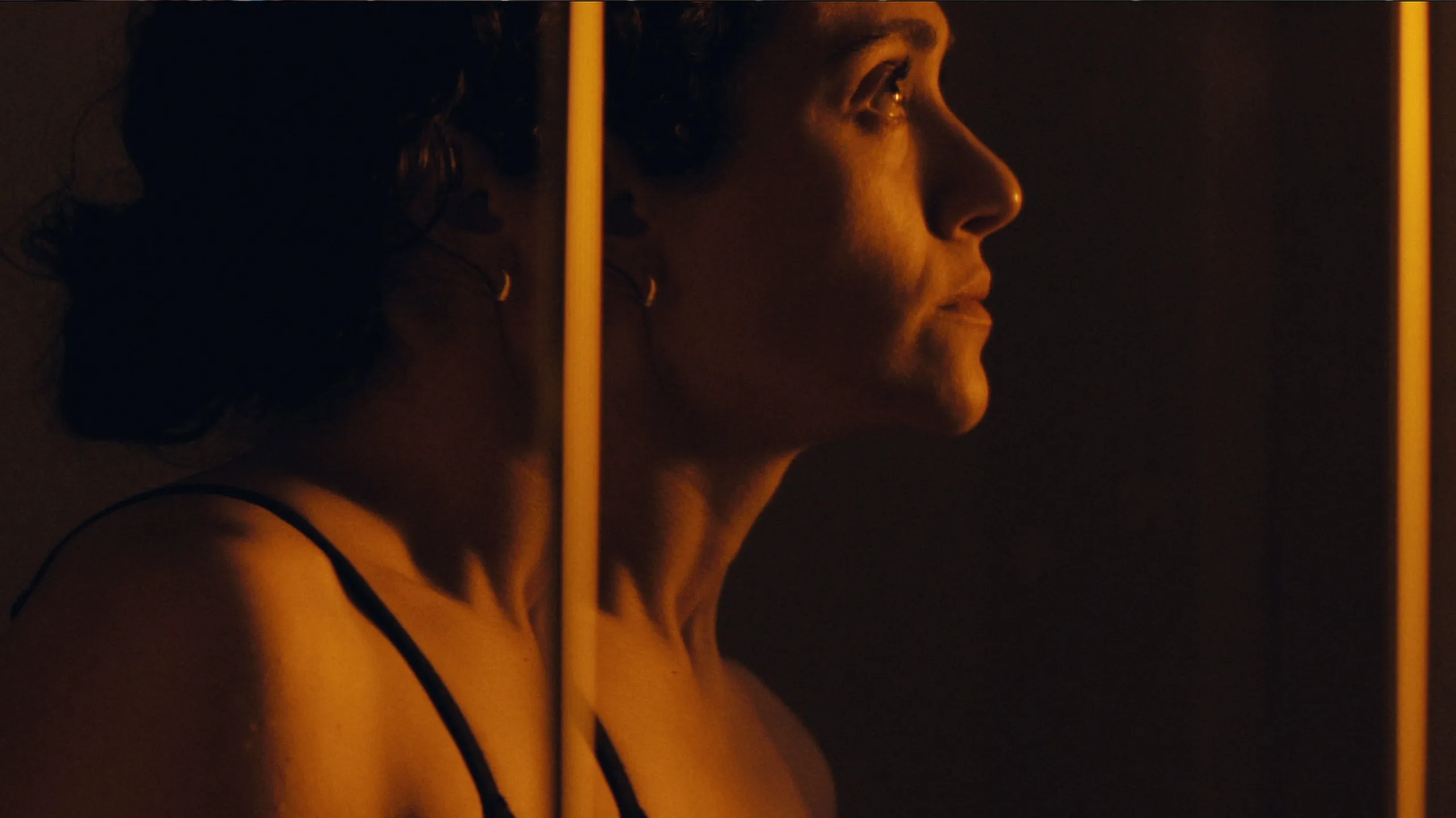 Alice stands beside a mirror, shown in profile under warm lighting.