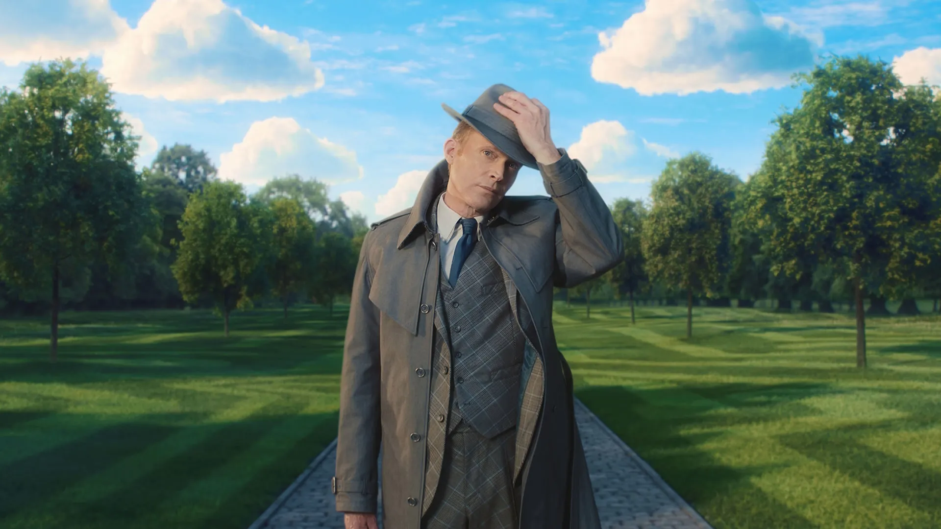 Vision in human disguise tipping his gray fedora while wearing a trench coat and a plaid three-piece suit, standing on a cobblestone path centered within a surreal, manicured park under a bright blue sky with puffy white clouds in Marvel Television's VisionQuest.