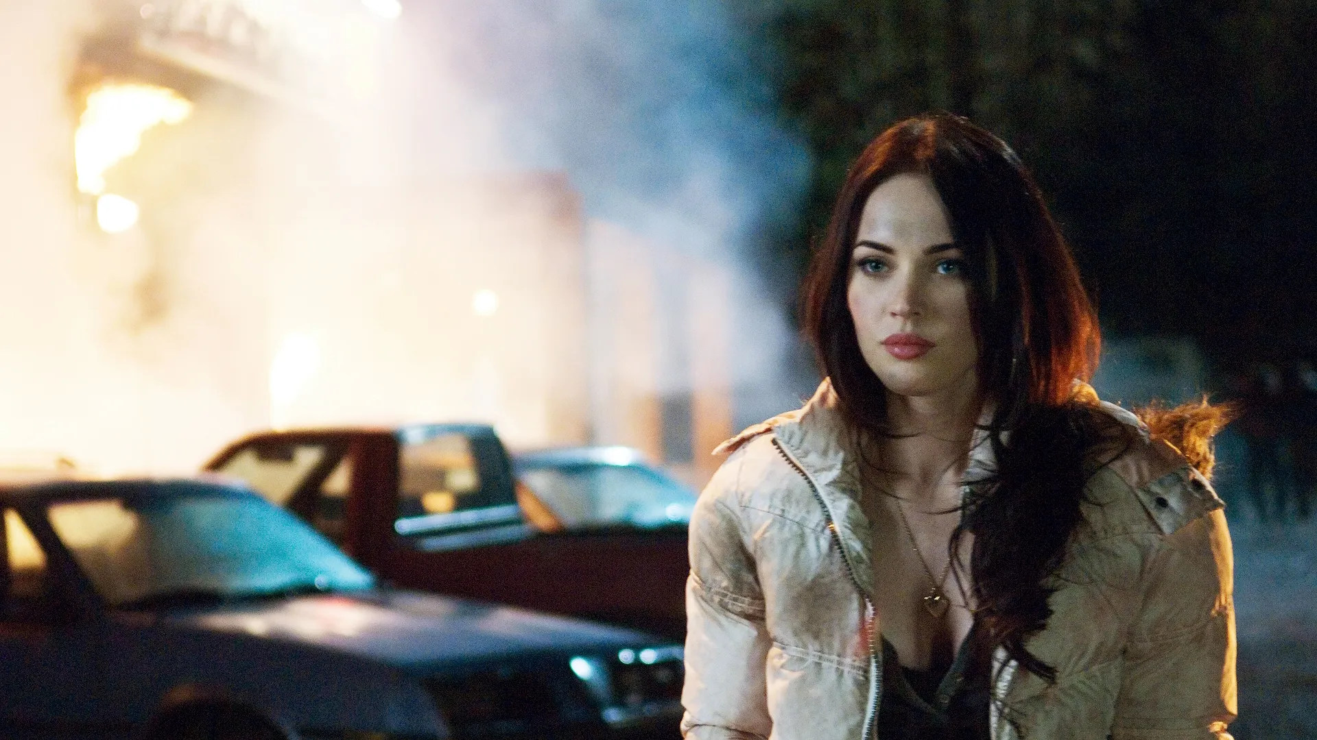 Megan Fox as Jennifer Check stands outside a burning building at night, wearing a dirty white puffer jacket, from the horror-comedy film Jennifer's Body.