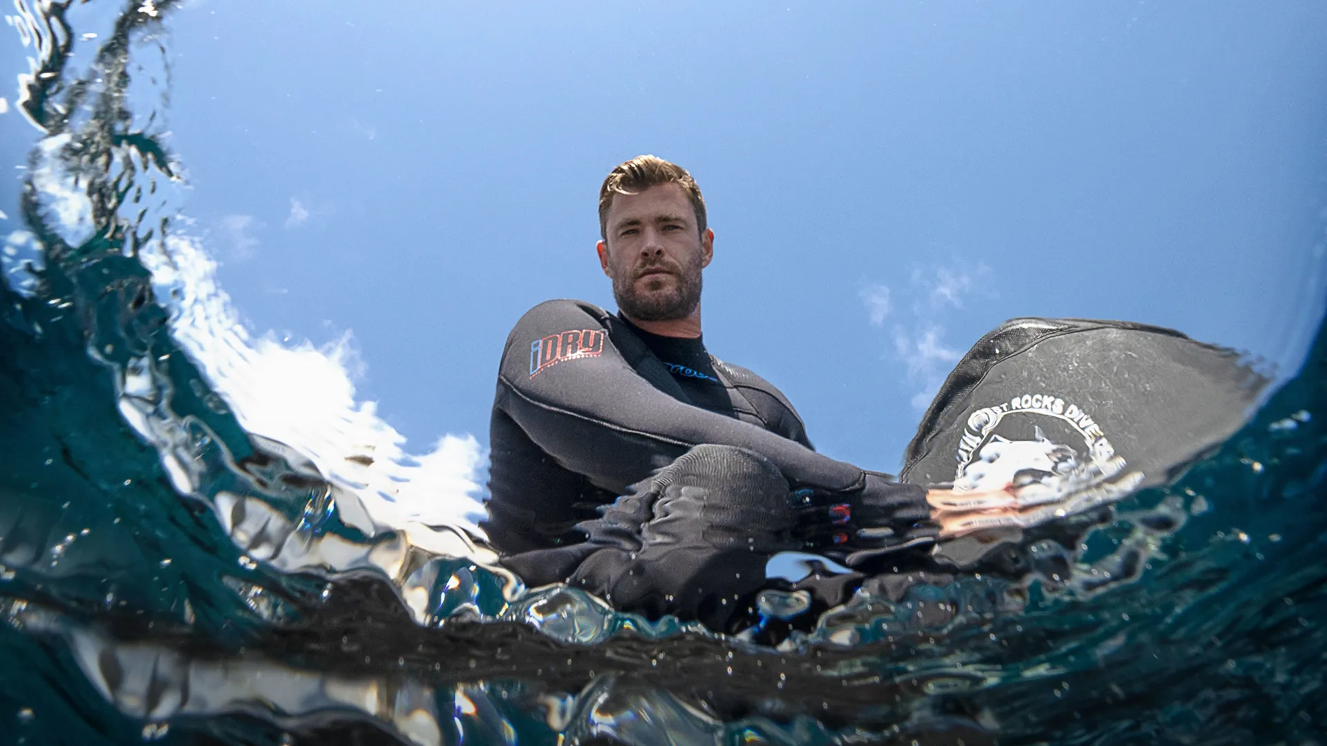Chris Hemsworth looking down at the water while wearing a wetsuit.