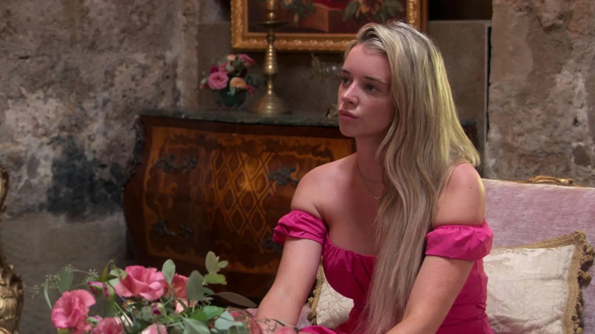 Hannah sits pensively on a pink armchair in a room decorated with bouquets of roses