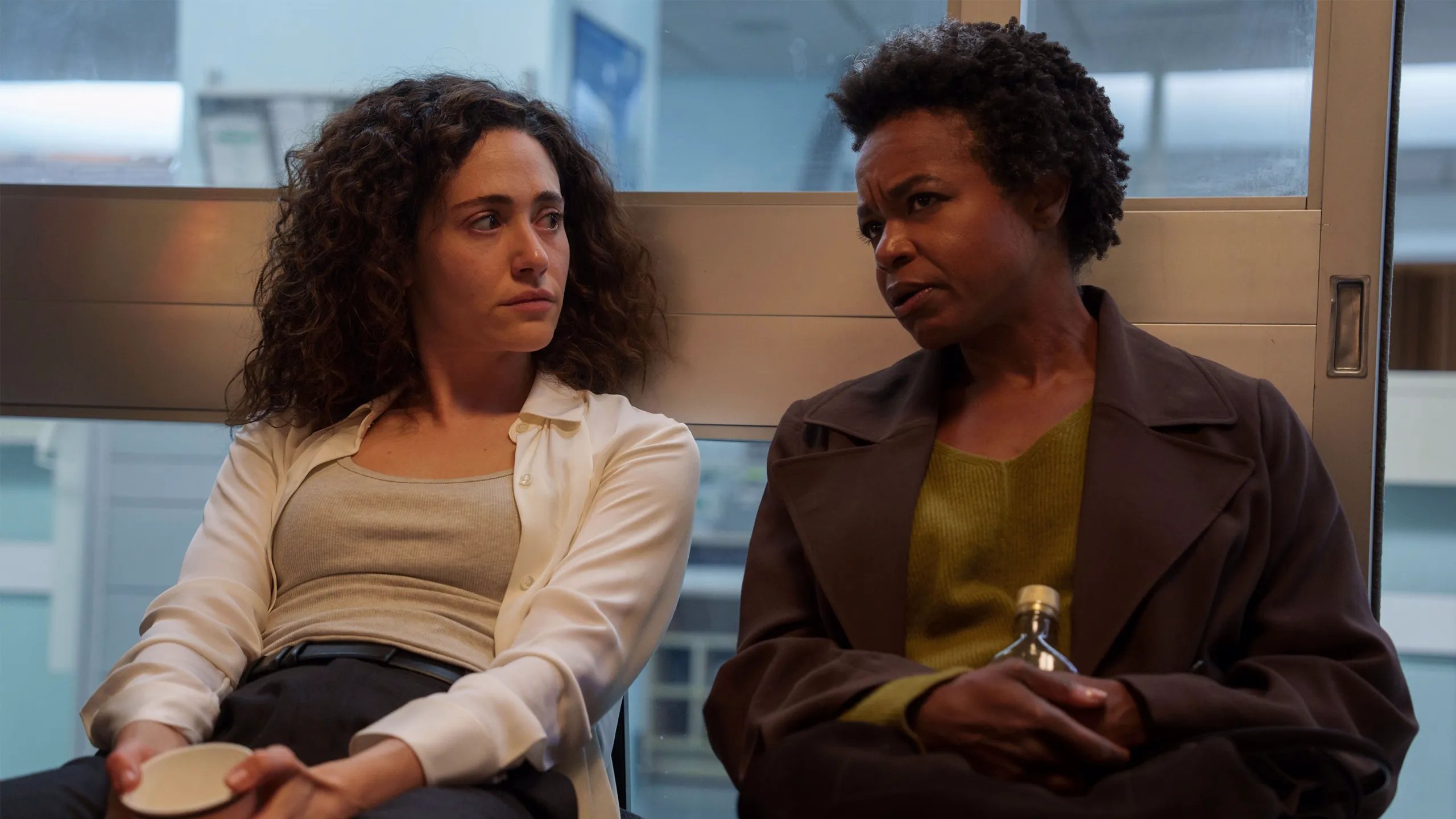 Alice and Nora sit together in a corridor talking.