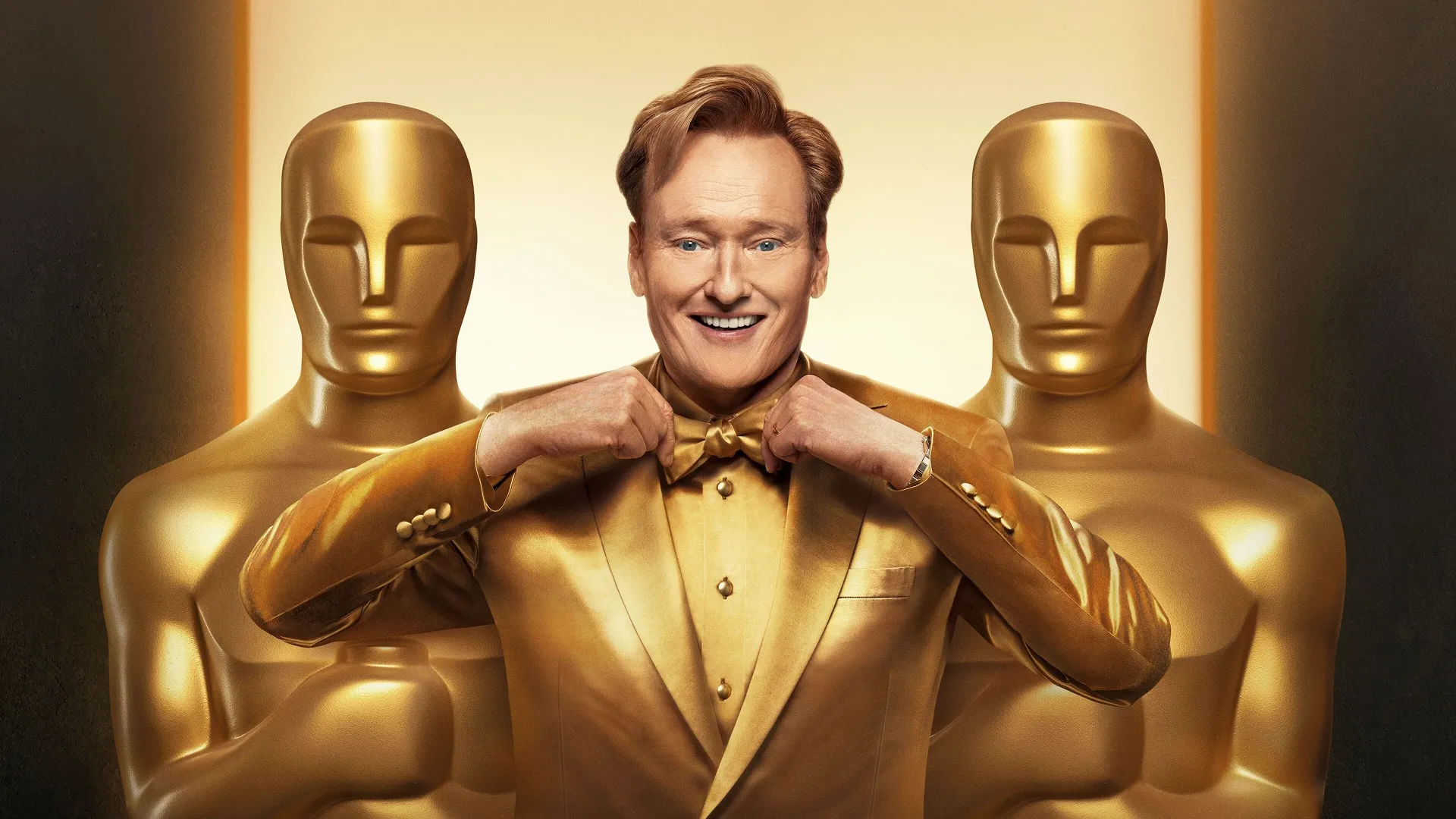 Oscars host Conan O'Brien in a gold suit stand's behind two human sized Oscar's trophies.