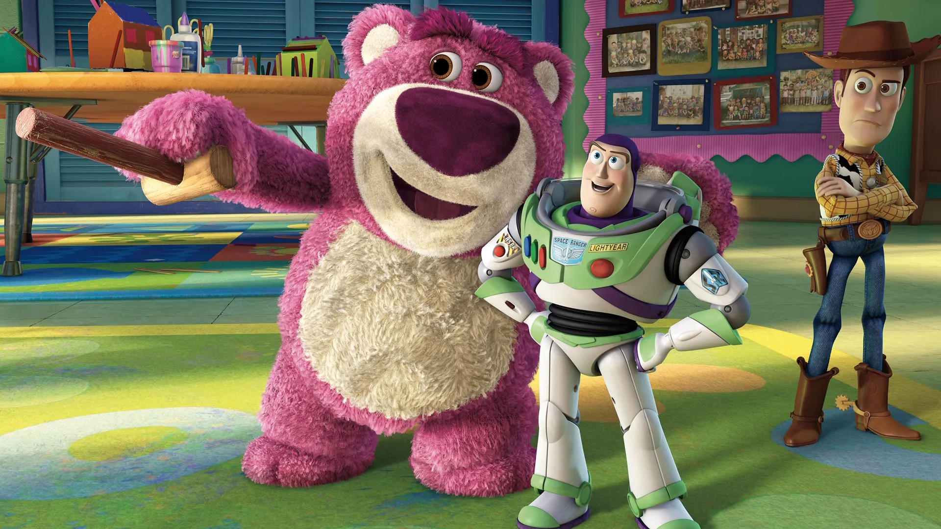 The large pink teddy bear Lotso smiles and gestures with a wooden cane next to an optimistic Buzz Lightyear, while a skeptical Woody stands in the background inside a colorful daycare classroom in a still from Toy Story 3.