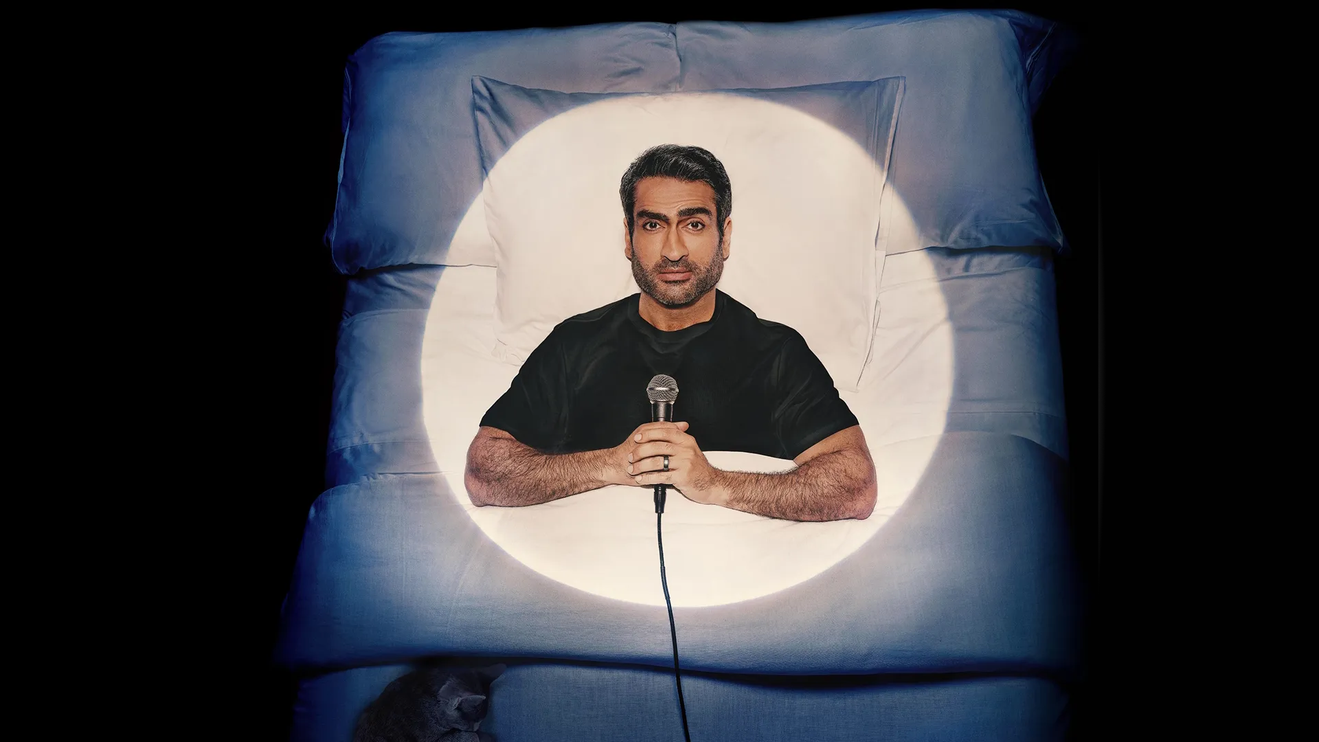 Comedian Kumail Nanjiani lying in a bed with a microphone in his hands.