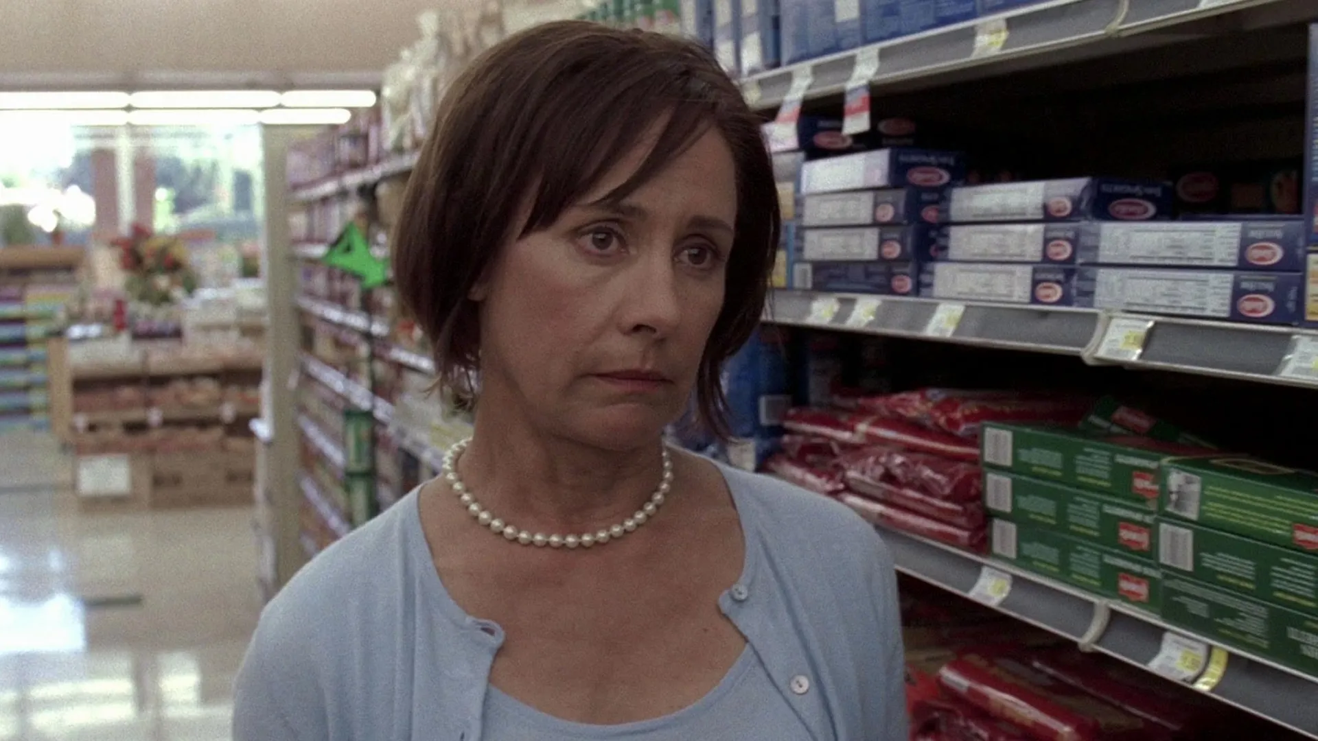 A somber Carolyn Bigsby (Laurie Metcalf) with short brown hair stands in a grocery store aisle filled with pasta boxes, wearing a pearl necklace and a light blue cardigan.