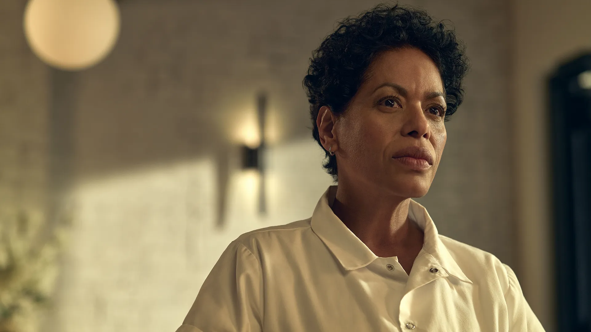 Close fechado de Tina Marrero (Liza Colón-Zayas), determinada, com cabelos curtos e cacheados, vestindo uma camisa branca de chef e olhando atentamente para fora do plano da câmera, dentro de um restaurante bem iluminado.