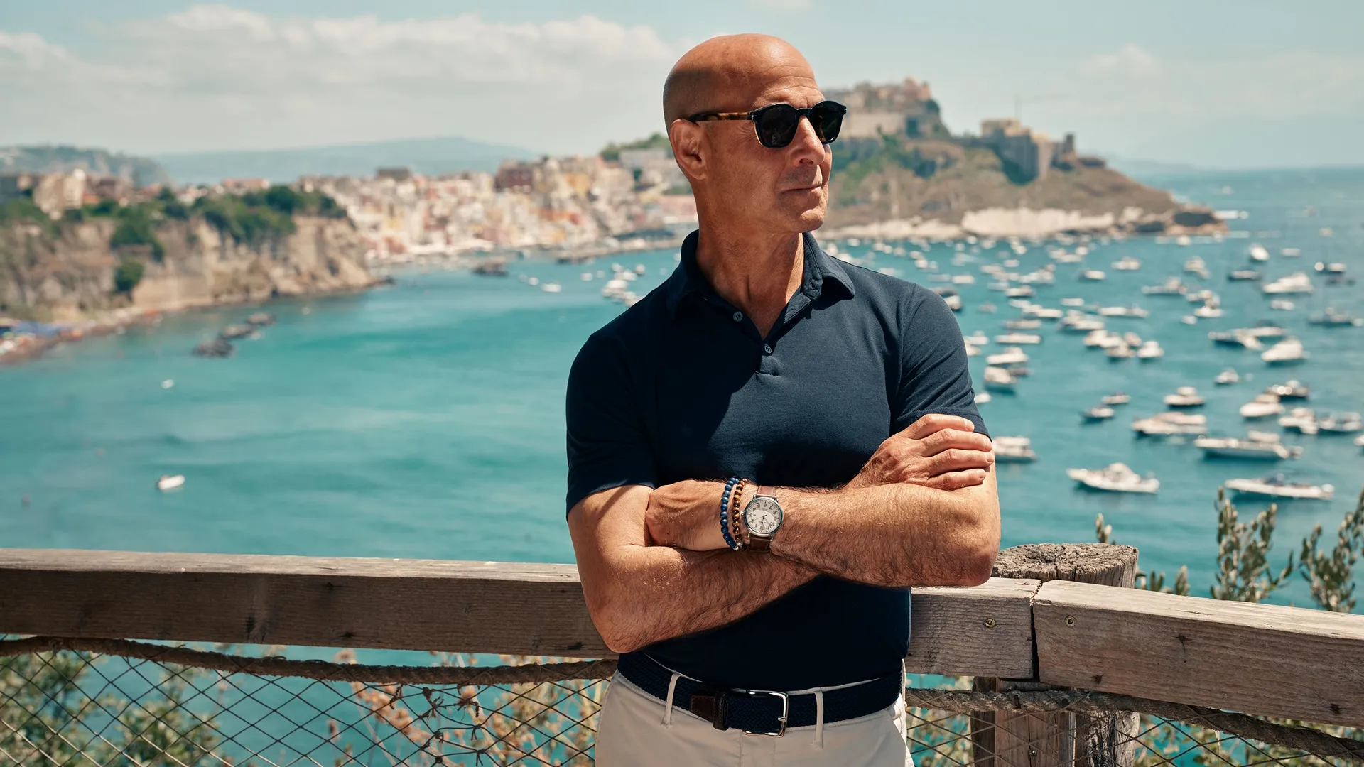 Stanley Tucci standing in front of an Italian coastal view, arms crossed and wearing sunglasses