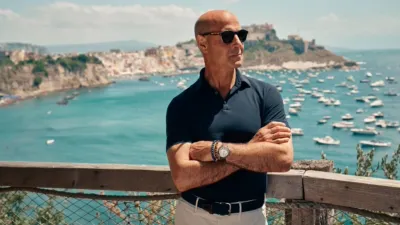 Stanley Tucci de pé diante de uma paisagem costeira italiana, com os braços cruzados e usando óculos de sol