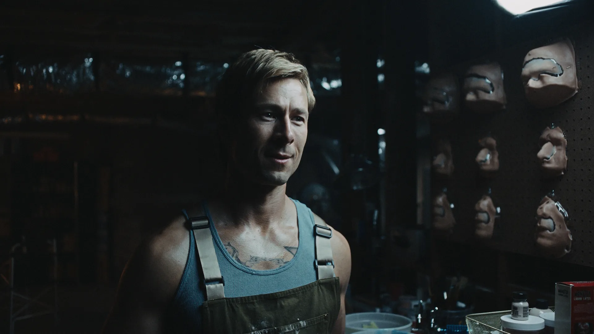 Um plano fechado mostra Russ Holliday (Glen Powell) sorrindo levemente em uma oficina mal iluminada, com uma regata cinza canelada e um macacão marrom, com moldes faciais protéticos em um painel perfurado ao fundo.