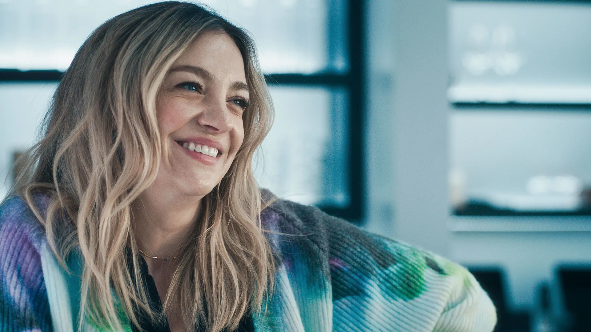 Close de Natalie "Sugar" Berzatto (Abby Elliott) sorrindo calorosamente enquanto olha para fora do enquadramento, vestindo um cardigã vibrante com decote em V e estampa tie-dye.
