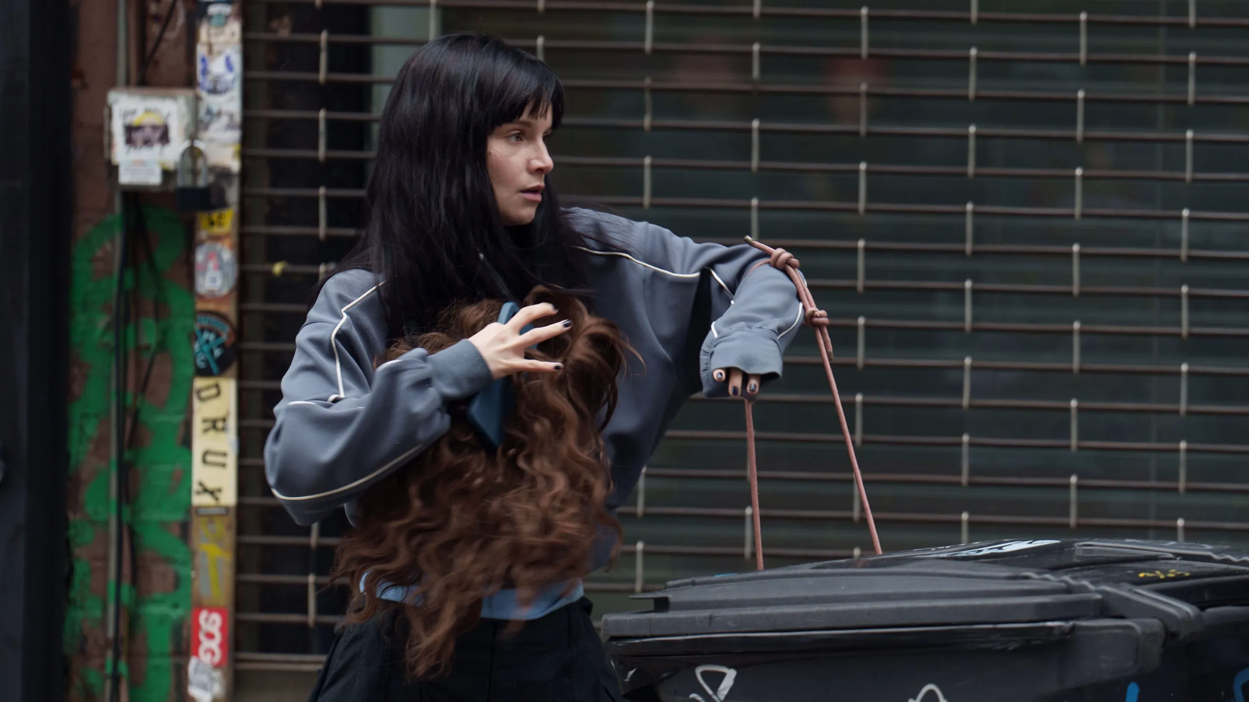 Catherine holds a long wig beside rubbish bins in a city street.