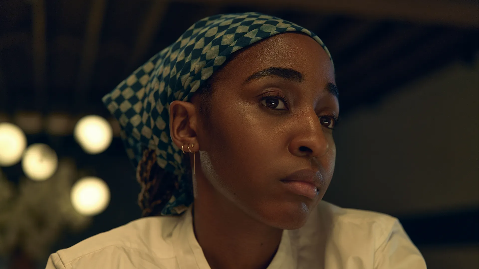 Um close mostra Sydney Adamu (Ayo Edebiri) concentrada, usando um uniforme branco de chef e um lenço na cabeça estampado verde e branco, olhando para o lado, em um restaurante.