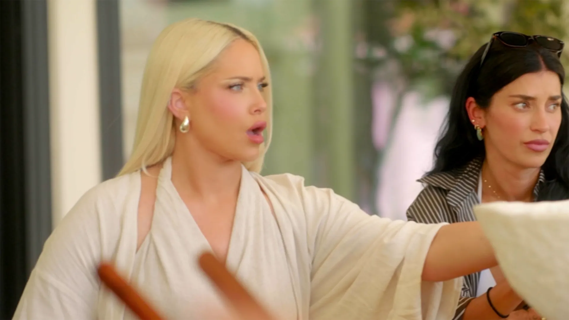 A woman with long blonde hair looks to the side with an open mouth while reaching her hand forward, next to a woman with long dark hair who is watching her intently.