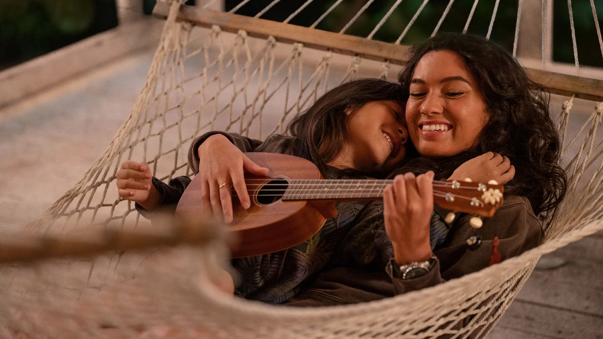 Lilo lies in a hammock with her sister, both smiling as Nani plays the ukelele.