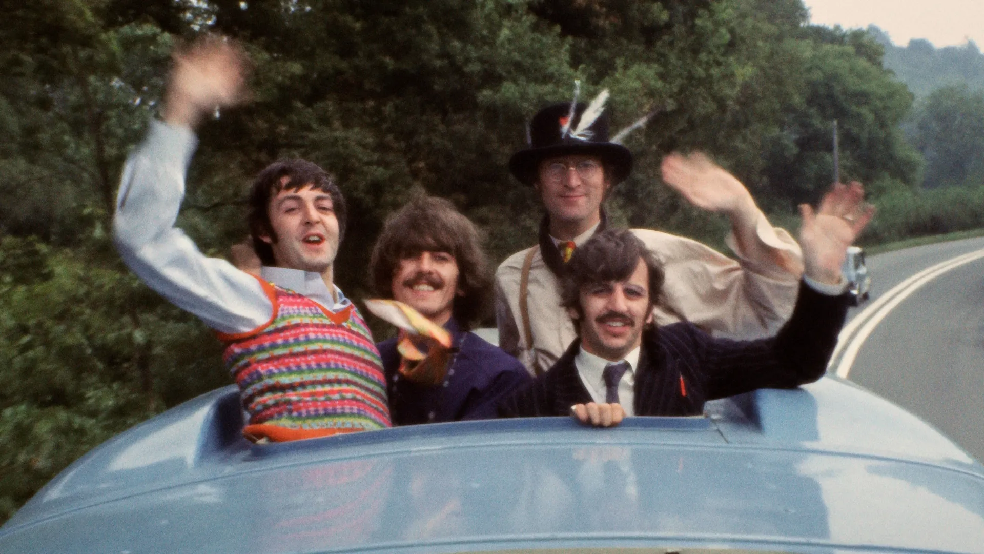 The Beatles (L-R: Paul McCartney, George Harrison, John Lennon, and Ringo Starr) waving from the sunroof of a blue car in their 1967 surrealist comedy film, Magical Mystery Tour.