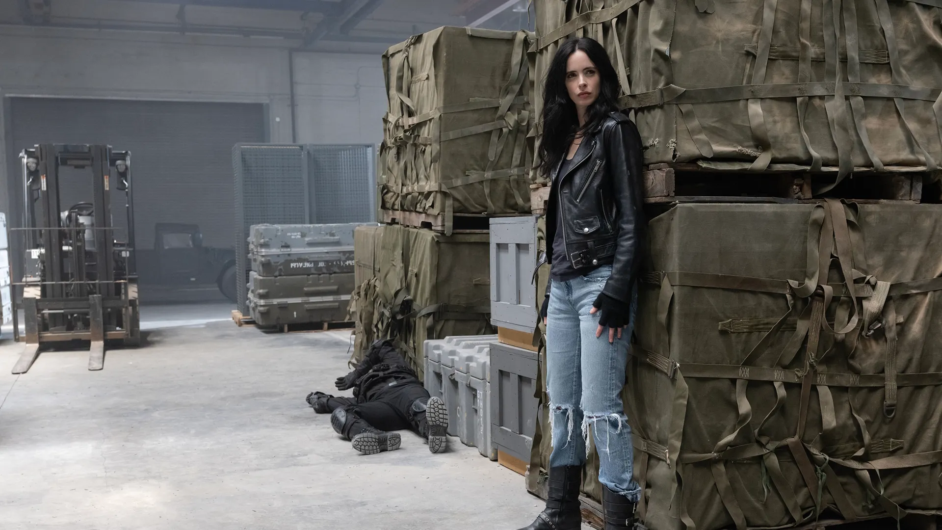 Jessica Jones (Krysten Ritter) hides behind crates of weapons in a warehouse.