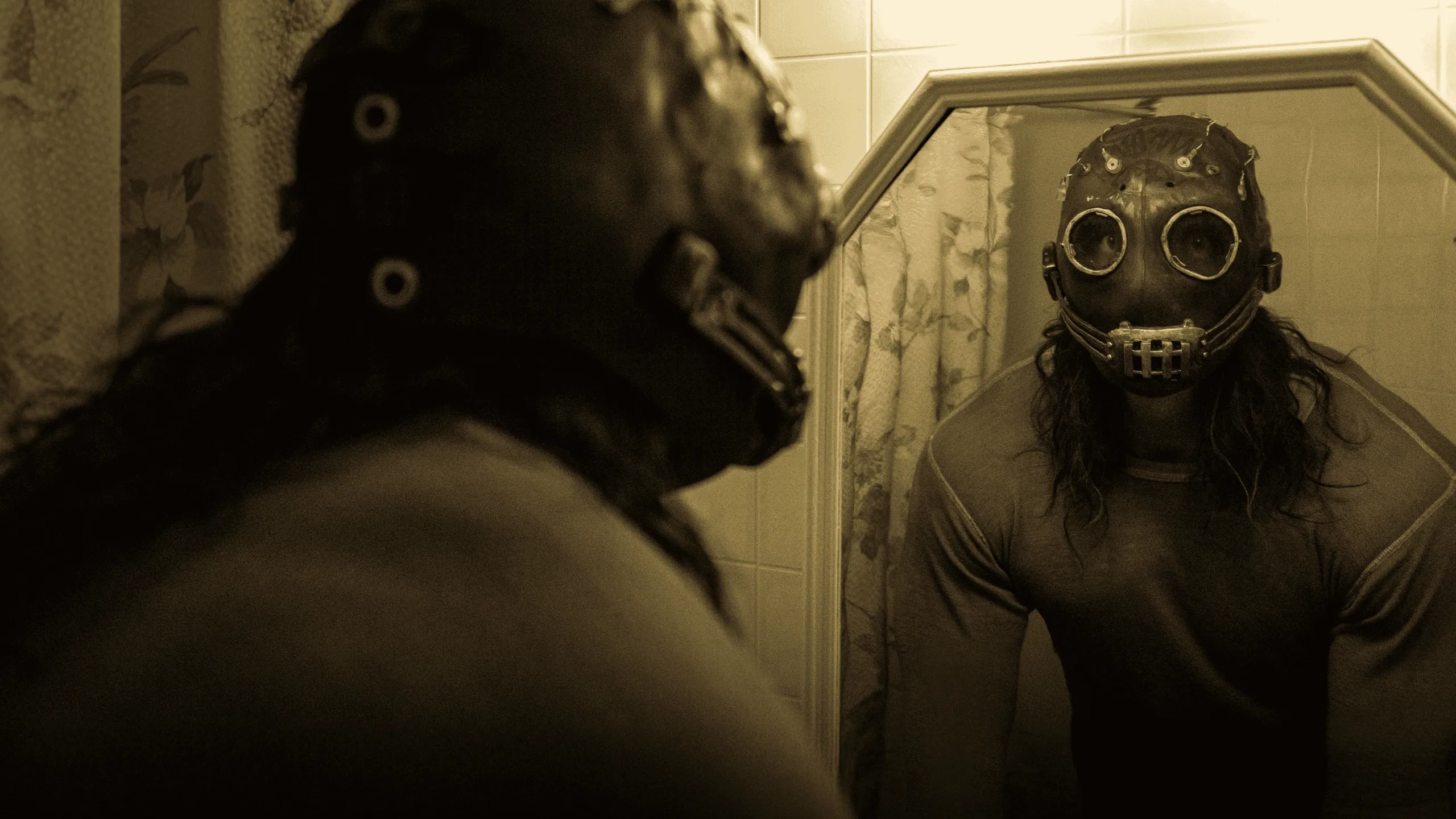 A close-up film still from the horror thriller Psycho Killer, showing the Satanic Slasher (James Preston Rogers) wearing a sinister, studded leather gas mask over long hair while staring intensely into a bathroom mirror.