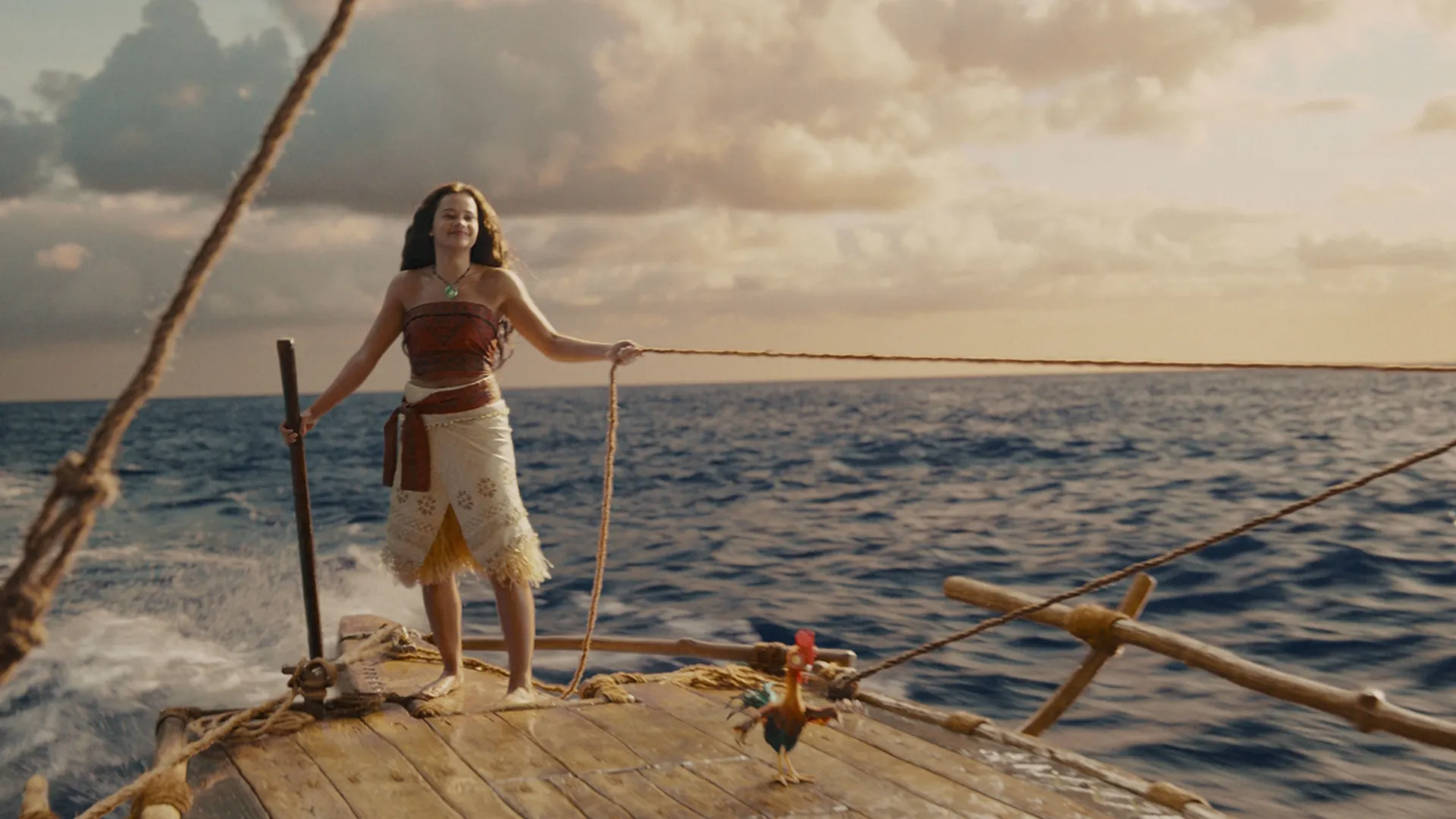 A live-action Moana stands smiling on the deck of her wooden canoe at sea, pulling a rope with Heihei the rooster nearby.