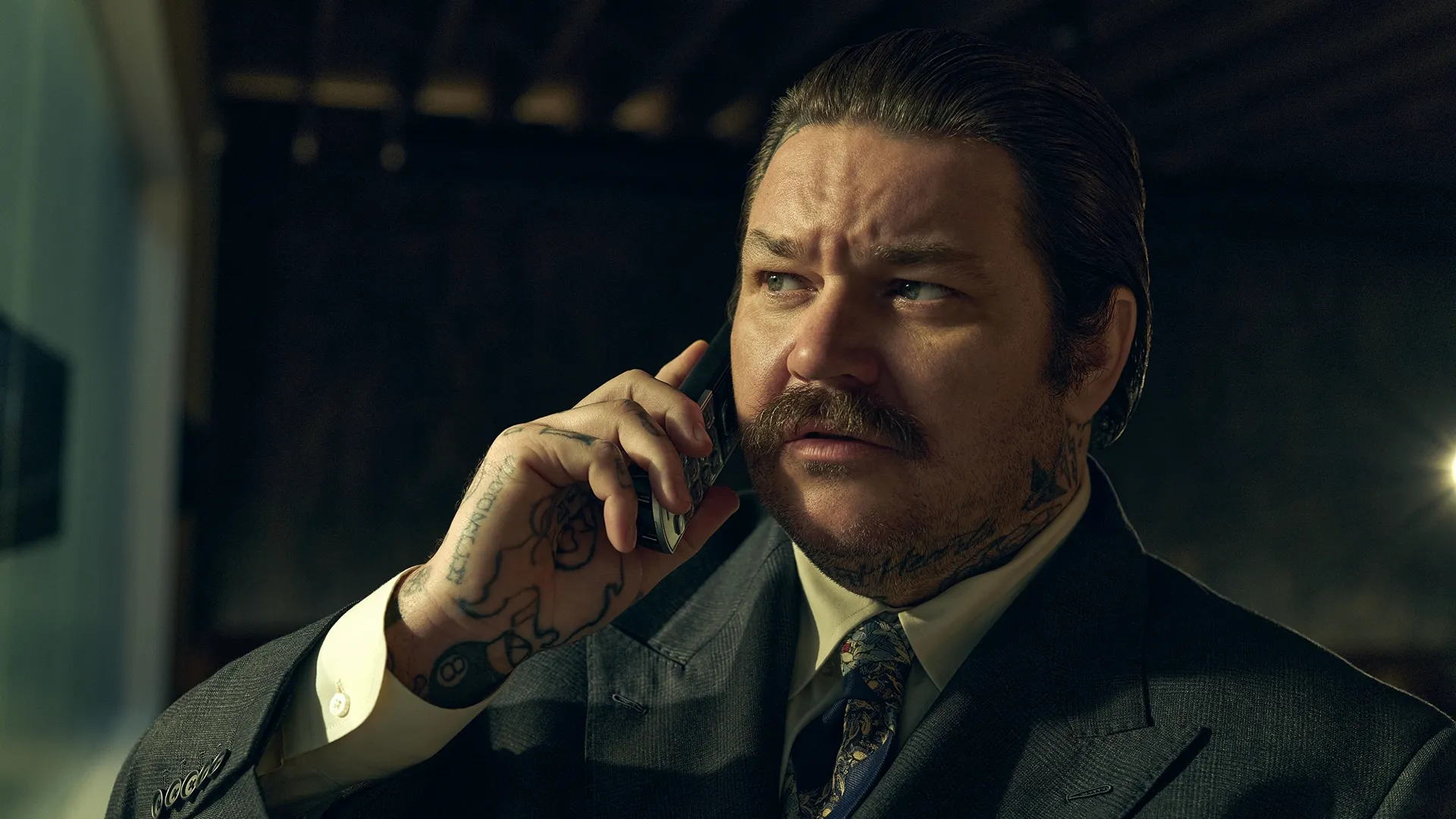 Close de Neil Fak (Matty Matheson) com cabelo penteado para trás e bigode, usando uma gravata estampada e um paletó cinza, enquanto segura um celular junto à orelha com a mão direita muito tatuada.