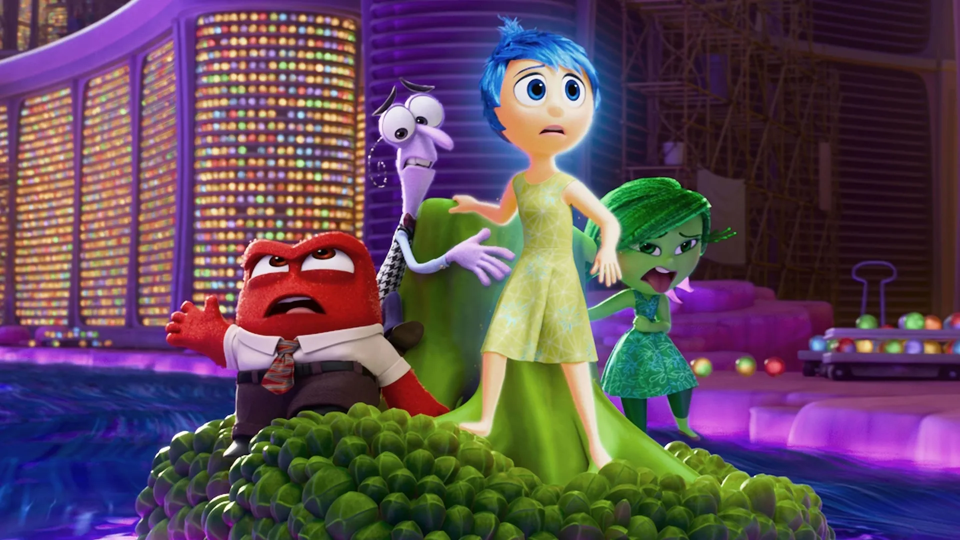 The bright blue-haired character Joy stands at the front of a floating green mass with a worried expression, while Anger, Fear, and Disgust react anxiously behind her against a vibrant backdrop of glowing, multi-colored memory shelves in a scene from the animated movie Inside Out 2.