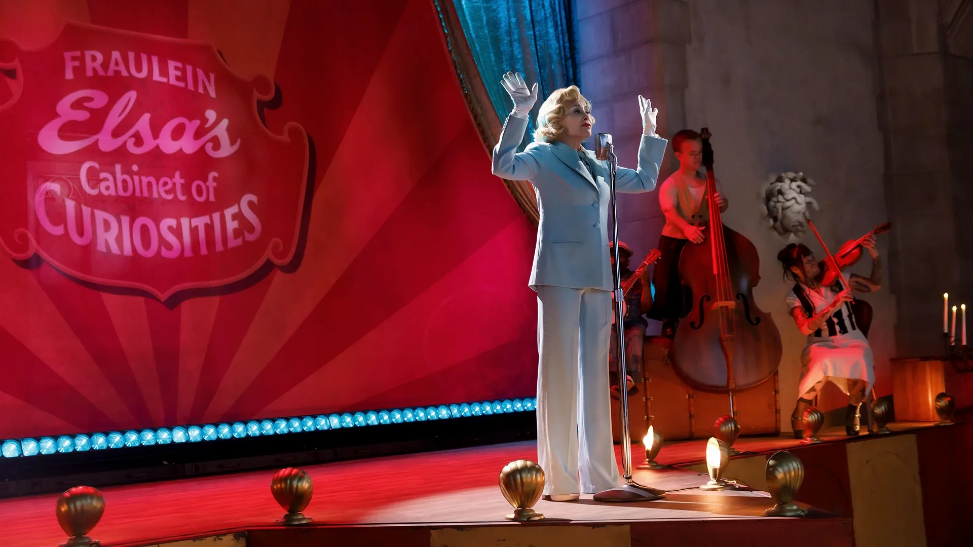 A blonde woman in a light blue pantsuit and white gloves sings into a vintage microphone on a stage featuring a sign for "Fraulein Elsa's Cabinet of Curiosities," accompanied by a band performing in the background.